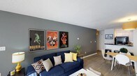 Located at 1700 Chapel Hill Rd in Durham, North Carolina, this single-floor residence offers a well-appointed and thoughtfully designed living space. The living area serves as the centerpiece of the home, featuring a striking dark gray accent wall that provides a bold backdrop for a curated gallery of vintage-style framed posters, adding a retro, artistic character to the room. Warm hardwood-style flooring runs throughout, complementing the contemporary layout and creating a cohesive, inviting atmosphere. The textured ceiling adds a classic touch, while recessed lighting and wall-mounted light fixtures provide warm, ambient illumination throughout the space. The open layout connects the living area to a hallway that leads further into the home, suggesting a practical and efficient floor plan. A glimpse of a dining area is visible in the background, hinting at a seamless flow between living and dining spaces. Overall, the space strikes a balance between modern design sensibilities and eclectic, personality-driven décor, making it a comfortable and visually engaging environment. Its single-floor layout ensures easy accessibility and a straightforward, connected living experience.