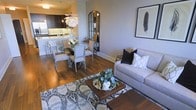 This single-floor residence showcases a thoughtfully designed open-concept layout, where the living, dining, and kitchen areas flow naturally into one another, creating a cohesive and functional living environment. Rich hardwood flooring extends throughout the space, adding warmth and continuity to the overall aesthetic.

The living area is anchored by a plush, neutral-toned sofa dressed with contrasting black and patterned cushions, complemented by a wooden coffee table resting atop a bold geometric area rug. A framed feather artwork adorns the light grey wall, adding an artistic focal point, while an arched mirror leaning nearby enhances the sense of depth and light in the room.

The dining area features a round glass-top table surrounded by elegantly upholstered chairs, illuminated by a striking white floral chandelier that serves as a decorative centerpiece. This area transitions seamlessly into the kitchen, which is equipped with dark cabinetry, stainless steel appliances, and a peninsula with bar seating, offering both style and practicality. The cohesive colour palette of soft greys, warm wood tones, and dark accents creates a sophisticated and inviting ambiance throughout.