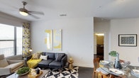 Located in Dallas, this single-floor residence offers a thoughtfully designed living space that blends modern aesthetics with everyday functionality. The open-concept layout seamlessly connects the living and dining areas, creating a cohesive and inviting environment. The living area features smooth, light-toned walls adorned with bold abstract artwork in yellow and white, adding a striking visual focal point. A stylish dark sectional sofa anchors the space, complemented by a geometric black-and-white area rug and a gold accent side table that adds a warm metallic touch. A ceiling fan ensures year-round comfort overhead. The dining area is positioned adjacent to the living space, furnished with a round glass-top table and mid-century modern wooden chairs, reinforcing the contemporary character of the home. Dark flooring runs throughout, providing a grounded contrast to the bright walls and lighter furnishings. A hallway visible from the main living area leads to additional rooms, with a glimpse of a well-appointed bathroom featuring dark cabinetry and warm lighting. The overall ambiance is clean, modern, and carefully curated, making this Dallas residence a polished and functional living space.