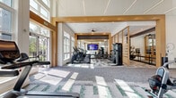Located at 4710 Mint Leaf Ln in Raleigh, NC, this single-floor fitness center is thoughtfully designed to provide a comprehensive wellness experience. The space is defined by soaring ceilings with warm wood-accented architectural framing, recessed and linear lighting, and an abundance of natural light streaming through large, multi-pane windows that line the perimeter walls. The patterned carpet tile flooring in shades of gray and green adds visual interest while providing a comfortable underfoot surface throughout the workout areas. The main fitness floor is generously proportioned and equipped with an extensive array of cardio machines, including treadmills, ellipticals, stair climbers, and stationary cycling bikes, complemented by cable strength machines, free weights, a rowing machine, and a wall-mounted display screen. Full-length mirrors and warm wood partition screens define a dedicated strength zone, enhancing both function and aesthetics. A stylish entryway features decorative wall art, a refreshment station, and a ceiling fan, creating a welcoming arrival experience. Direct access to an outdoor area provides artificial turf for recreational activities and additional outdoor fitness equipment, all set against a backdrop of lush trees and well-maintained landscaping. The exterior presents a clean, contemporary facade with large glass windows and manicured grounds.