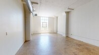 Located in the heart of downtown Los Angeles at 443 San Pedro St, this single-floor unit offers an open, loft-inspired layout defined by a blend of industrial and contemporary design elements. Polished concrete floors extend throughout the space, lending a raw yet refined aesthetic that anchors the overall ambiance.

The main living area is expansive and airy, characterized by white-painted walls, exposed cylindrical ductwork running along the ceiling, and large multi-pane windows that invite ample natural light. Structural columns and ceiling beams contribute to the industrial character of the space. Track lighting and linear LED fixtures provide practical illumination across the open floor plan.

The kitchen is outfitted with light wood-finish cabinetry, dark countertops, a gold-toned undermount sink with a matte black faucet, a subway-style tile backsplash, and stainless steel appliances including a range, microwave, refrigerator, and dishwasher. A set of black-framed interior doors adds a modern, graphic contrast to the white walls. The bathroom features white mosaic tile flooring, a fully tiled walk-in shower, and accessibility-friendly grab bars with matte black hardware. An additional room with exposed ductwork and a warm wood entry door provides flexible use as a bedroom, office, or studio space.