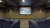 This dedicated movie theater is thoughtfully designed to deliver a true cinema experience in an intimate setting. At the front of the room, a large projection screen dominates the wall, supported by a ceiling-mounted projector and a surround sound speaker system positioned throughout the space for immersive audio. The seating area features multiple rows of generously sized, navy blue upholstered recliners, each equipped with built-in cup holders and accented with plaid throw pillows, offering both comfort and style. The rows are arranged to ensure clear sightlines to the screen from every seat. Underfoot, a bold striped carpet in neutral tones adds visual interest and contributes to the theater's polished aesthetic. The dark ceiling and warm-toned walls create a focused, atmospheric ambiance ideal for movie viewing. Wall-mounted sconces provide subtle ambient lighting between features. Classic framed movie posters — including iconic titles such as Dracula, Gone with the Wind, and Casablanca — line the side walls, reinforcing the cinematic theme. A built-in snack counter with dark cabinetry and a light countertop is positioned along one wall, providing a convenient space for refreshments, completing the full movie-going experience.