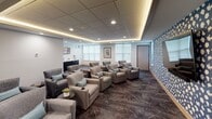 This dedicated media lounge offers a refined and comfortable group viewing experience within a single, thoughtfully designed room. The space is arranged with rows of plush, upholstered swivel armchairs in a warm gray fabric, each accented with teal throw pillows and equipped with built-in cup holders for added convenience. The seating is oriented toward a large wall-mounted flat-screen television, anchored against a striking bold blue-and-white patterned accent wall that serves as a dramatic focal point.

The ceiling features a coffered drop-tile design with integrated ambient LED cove lighting that casts a warm, golden glow around the room's perimeter, complementing the recessed downlights for a layered, cinematic atmosphere. The flooring consists of richly patterned, dark-toned carpet that absorbs sound and enhances the room's acoustic qualities.

Natural light is introduced through a row of windows along one wall, fitted with light-diffusing shades to allow brightness control during screenings. Framed artwork adorns the walls, adding a polished, hospitality-inspired aesthetic. A small credenza or service counter is visible in one corner, suggesting optional refreshment service. The overall design balances modern elegance with functional comfort, making this space ideal for movie screenings, presentations, or group entertainment.