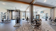 Located in Chantilly, VA, this well-appointed fitness center offers a generous, single-floor layout designed to accommodate a full range of workout needs. The space is thoughtfully divided into distinct training zones, with durable rubber flooring in the strength and free weights areas transitioning to patterned carpet tile throughout the cardio and functional training sections. Exposed wood-toned beams and structural columns add architectural character, while a large ceiling fan ensures comfortable air circulation throughout. Recessed lighting keeps the space bright and well-illuminated at all times. An impressive array of Matrix and cable-based strength machines occupies the central floor, complemented by adjustable benches, a multi-station cable tower, and a dedicated free weights area with a full dumbbell rack. The cardio zone features treadmills, stationary bikes — including Peloton cycles — ellipticals, and rowing machines, all positioned to take advantage of floor-to-ceiling windows and glass doors that invite abundant natural light and provide views of the surrounding outdoor areas. Wall-mounted mirrors expand the sense of space and assist with form during training. A medicine ball and TRX suspension training station rounds out the functional fitness offerings. Wall-mounted televisions enhance the overall exercise experience in this modern, comprehensively equipped facility.