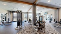 Located in Chantilly, VA, this well-appointed fitness center offers a generous, single-floor layout designed to accommodate a full range of workout needs. The space is thoughtfully divided into distinct training zones, with durable rubber flooring in the strength and free weights areas transitioning to patterned carpet tile throughout the cardio and functional training sections. Exposed wood-toned beams and structural columns add architectural character, while a large ceiling fan ensures comfortable air circulation throughout. Recessed lighting keeps the space bright and well-illuminated at all times. An impressive array of Matrix and cable-based strength machines occupies the central floor, complemented by adjustable benches, a multi-station cable tower, and a dedicated free weights area with a full dumbbell rack. The cardio zone features treadmills, stationary bikes — including Peloton cycles — ellipticals, and rowing machines, all positioned to take advantage of floor-to-ceiling windows and glass doors that invite abundant natural light and provide views of the surrounding outdoor areas. Wall-mounted mirrors expand the sense of space and assist with form during training. A medicine ball and TRX suspension training station rounds out the functional fitness offerings. Wall-mounted televisions enhance the overall exercise experience in this modern, comprehensively equipped facility.