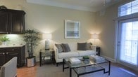 Located at 250 Liberty Street in Columbus, Ohio, this single-floor space offers a refined and welcoming atmosphere defined by clean lines and a carefully curated aesthetic. The living area serves as the centerpiece of the space, showcasing a neutral color palette of warm beige and soft gray tones that create a calm, sophisticated ambiance throughout. The room is anchored by a classic white upholstered sofa accented with layered decorative pillows, complemented by sleek glass-top coffee and side tables with dark metal frames that lend a contemporary edge. Warm ambient lighting from stylish table lamps on each side of the seating area creates an inviting glow, while a framed artwork above the sofa adds a tasteful focal point to the wall. A potted indoor plant in the corner introduces a natural, organic element, softening the modern furnishings and bringing life to the space. The hardwood flooring beneath a textured area rug adds warmth and depth, contributing to the overall sense of comfort and quality. Crown molding details along the ceiling add a refined architectural touch, enhancing the polished feel of the interior. Overall, this space balances modern design sensibilities with a cozy, livable charm well-suited to its Columbus, OH location.