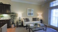 Located at 250 Liberty Street in Columbus, Ohio, this single-floor space offers a refined and welcoming atmosphere defined by clean lines and a carefully curated aesthetic. The living area serves as the centerpiece of the space, showcasing a neutral color palette of warm beige and soft gray tones that create a calm, sophisticated ambiance throughout. The room is anchored by a classic white upholstered sofa accented with layered decorative pillows, complemented by sleek glass-top coffee and side tables with dark metal frames that lend a contemporary edge. Warm ambient lighting from stylish table lamps on each side of the seating area creates an inviting glow, while a framed artwork above the sofa adds a tasteful focal point to the wall. A potted indoor plant in the corner introduces a natural, organic element, softening the modern furnishings and bringing life to the space. The hardwood flooring beneath a textured area rug adds warmth and depth, contributing to the overall sense of comfort and quality. Crown molding details along the ceiling add a refined architectural touch, enhancing the polished feel of the interior. Overall, this space balances modern design sensibilities with a cozy, livable charm well-suited to its Columbus, OH location.