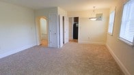 Located at 210 C, this single-floor space offers a practical and comfortable layout with a cohesive design throughout. The main living area is finished with plush neutral-toned carpeting that adds warmth underfoot and flows seamlessly through the space. Soft, light green walls create a calm and welcoming ambiance, complemented by clean white trim and baseboards that frame each room neatly.

A distinctive arched passageway on one side of the living area adds an architectural touch, leading into an adjoining carpeted room that provides additional living space. A standard hinged door nearby opens to what appears to be a closet or utility area, offering convenient built-in storage. On the opposite side, an open doorway provides a direct sightline into the kitchen, where dark cabinetry and a black refrigerator are visible, hinting at a well-equipped cooking space.

A pendant light fixture hangs in the dining area, providing focused overhead lighting and serving as a subtle design accent. The overall layout feels open and connected, with clearly defined zones for living, dining, and access to other areas of the unit, making the space functional and easy to navigate.