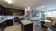 This well-appointed single-floor residence offers a thoughtfully designed open-concept layout that seamlessly connects the kitchen, dining, and living areas. The kitchen is a standout feature, showcasing rich dark cabinetry, granite countertops, a central island with pendant lighting overhead, and a full suite of stainless steel appliances including a range, microwave, dishwasher, and refrigerator. A clean white subway tile backsplash adds a modern touch. Light hardwood-style flooring flows through the main living areas, enhancing the bright and airy feel. The dining area accommodates a round table and chairs, positioned conveniently between the kitchen and the living room. The living room benefits from large windows that invite ample natural light and features a ceiling fan for added comfort. Two bedrooms offer plush carpeting and generous natural light; one includes a walk-in closet with direct access to an ensuite bathroom. The primary bathroom features a dual-sink vanity with granite countertops, dark cabinetry, a large mirror with overhead lighting, and a full tub-shower combination. A second full bathroom provides additional convenience, also finished with matching granite and dark cabinetry. The overall design presents a cohesive, contemporary aesthetic throughout.
