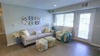 This single-floor space features a thoughtfully designed living room that blends comfort and style. The room is finished with light hardwood flooring and soft sage-gray walls, creating a calm, airy atmosphere. A large window fitted with white blinds allows ample natural light to fill the space, offering a view of lush greenery outside. Recessed ceiling lighting provides additional illumination, enhancing the room's bright and welcoming feel. The living area is furnished with a generously sized neutral-toned sofa accented with yellow and white decorative pillows, complemented by a matching armchair draped with a teal throw blanket. Two patterned ottomans in shades of teal, yellow, and white serve as both functional seating and decorative focal points. A side table with a classic lamp and decorative accent adds a refined touch. The wall features an eclectic gallery arrangement of ornate and geometric frames in varying shapes, adding visual interest. An HVAC vent is integrated into the upper wall, ensuring year-round comfort. Overall, the space exudes a relaxed yet polished aesthetic.