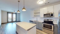 This single-floor space features a thoughtfully designed open-concept layout that seamlessly connects the kitchen and living areas. The kitchen is a standout feature, showcasing a two-tone cabinet design with white upper cabinetry and gray lower cabinets, complemented by a classic white subway tile backsplash. A center island with a crisp white countertop serves as a functional focal point, illuminated overhead by a pair of brass dome pendant lights that add a warm, contemporary aesthetic. Stainless steel appliances, including a range, microwave, dishwasher, and French door refrigerator, complete the well-equipped cooking space. Light wood-look flooring runs throughout the main living areas, contributing to a bright and airy atmosphere. The bedroom offers plush carpeting underfoot and features a generously sized window that allows natural light to flow in while providing views of the surrounding urban landscape, along with a ceiling fan for added comfort. The bathroom is finished with a granite-topped vanity, an undermount sink, and a walk-in shower, offering a clean and functional design. Overall, the space presents a modern, cohesive aesthetic with quality finishes throughout.
