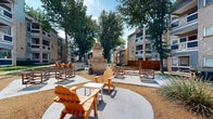 Located at 6700 Sandshell Blvd in Fort Worth, Texas, this multi-building residential community offers a well-designed outdoor living environment that encourages connection and relaxation. The grounds are anchored by a striking stone outdoor fireplace adorned with a Texas star emblem, surrounded by Adirondack chairs, wooden benches, and latticed seating clusters arranged across concrete pads and gravel gathering areas — ideal for socializing under the open sky.

Mature trees shade the winding concrete pathways that connect the various buildings and amenity areas throughout the property. The multi-story buildings feature a combination of stone and blue lap siding exteriors, with private balconies on upper floors offering views of the landscaped grounds below. Manicured shrubbery, native plantings, and a checkerboard stepping-stone lawn pattern add visual interest to the courtyard spaces.

A garage space on the property has been thoughtfully converted into a versatile community room, featuring exposed wood truss ceilings with track lighting, pendant fixtures, built-in bench seating with colorful cushions, and polished concrete floors — creating a lively, functional gathering space. A swimming pool area is also visible among the amenities. The overall atmosphere is warm, community-oriented, and distinctly Texan in character.