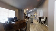 This single-floor office space offers a well-appointed and functional environment designed to balance professional utility with a welcoming atmosphere. The open floor plan allows for a clear sightline across the main areas, creating a sense of spaciousness throughout.

The primary workspace features a substantial dark wood executive desk paired with leather seating, positioned near a window fitted with horizontal blinds that allow for controlled natural light. A striking stacked stone fireplace serves as a focal architectural centerpiece, adding warmth and character to the interior. A decorative round metal wall accent adorns the fireplace surround, contributing a touch of style to the space.

Vaulted ceilings rise toward the rear of the space, accentuated by recessed and track lighting that ensures even, well-distributed illumination throughout. Light wood-tone laminate flooring runs continuously through all areas, tying the space together cohesively.

A casual lounge area features upholstered seating, providing a comfortable setting for informal meetings or waiting. Toward the back, a kitchenette area equipped with a microwave and cabinetry offers practical amenities for occupants. A high-top bistro table with stools adds a versatile gathering spot, rounding out the functional and thoughtfully arranged layout of this professional space.