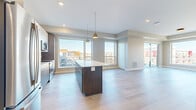 This contemporary single-floor residence offers a thoughtfully designed open-concept layout with clean lines and modern finishes throughout. The kitchen serves as the centerpiece of the living space, featuring a large dark-cabinetry island topped with a white quartz countertop, stainless steel appliances including a French-door refrigerator, microwave, and range, and stylish pendant lighting overhead. Light wood-tone flooring extends through the main living areas, enhancing the bright, airy atmosphere created by an array of large windows that frame views of the surrounding urban landscape.

The bedrooms are carpeted for comfort and each benefit from generous windows that bring in abundant natural light and city views. A spacious walk-in closet with built-in shelving and hanging rods provides ample storage. The bathroom is finished with neutral tile in the shower-tub combo, a wood-grain vanity with white countertop, and a framed mirror, offering a clean and functional design. An in-unit stacked Whirlpool washer and dryer adds everyday convenience. Additional rooms with light wood flooring provide flexible living or office space. The overall aesthetic is modern and neutral, making it a move-in ready urban residence.