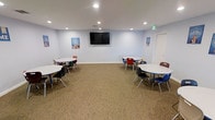 This single-floor space is designed as a functional and inviting group learning or activity room. The open layout is organized around several round tables, each surrounded by colorful chairs in red, blue, black, and green — an arrangement that encourages collaborative interaction and group work. The room's light blue walls create a calm, welcoming atmosphere, complemented by neutral carpeted flooring that adds warmth and acoustic comfort throughout the space.

Recessed ceiling lighting provides consistent, even illumination across the entire room, ensuring a well-lit environment suitable for focused activities. A large wall-mounted flat-screen display serves as a central visual focal point, ideal for presentations, instructional content, or group viewing. Educational posters are displayed on the walls, reinforcing the room's purpose as a structured learning environment.

The spacious floor plan allows for comfortable movement between seating areas and accommodates multiple small groups simultaneously. A clearly marked emergency exit is visible, and a door provides access to adjacent areas of the facility. The overall design prioritizes functionality, safety, and an engaging atmosphere well-suited for educational programs, workshops, tutoring sessions, or community group activities.