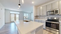 This well-designed single-floor residence offers a clean, contemporary aesthetic throughout, with a cohesive neutral palette and quality finishes at every turn. The open-concept kitchen serves as the heart of the home, showcasing grey shaker-style cabinetry, a tasteful stone tile backsplash, and a full suite of stainless steel appliances including a range, microwave, and French door refrigerator. A generous center island with a white quartz countertop and an undermount sink provides both additional prep space and a natural gathering point. Stylish pendant lighting hangs above the island, complementing the recessed lighting throughout. Light-toned hardwood flooring flows seamlessly from the kitchen into the open living area, which is bathed in natural light from large windows and a set of French doors that open to an outdoor space. A ceiling fan adds comfort to both the living and kitchen areas. The bedrooms feature plush carpeting and windows with views of surrounding greenery, while ceiling fans provide year-round comfort. Two full bathrooms are finished with white quartz countertops, grey cabinetry, large mirrors, and tiled tub-shower combinations, maintaining the home's consistent modern style. Overall, the space exudes a bright, airy ambiance with thoughtful design details throughout.