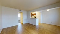 This single-floor space offers a straightforward, functional layout centered around an open living area finished with warm-toned hardwood flooring that runs throughout. The white walls and ceiling contribute to a bright, clean aesthetic, while a ceiling vent ensures comfortable climate control year-round.

A defining architectural feature is the pass-through kitchen counter, which connects the kitchen to the main living space. This cutout design allows visibility into the kitchen from the living area, promoting an open feel without fully removing the boundary between the two spaces. Inside the kitchen, warm-toned wood cabinetry is visible, adding a natural contrast to the otherwise neutral color palette.

A doorway off the main living area leads to an additional room, which appears to feature carpeted flooring and receives natural light through a window, offering a quieter, separate zone within the unit. The transition between rooms is clean and unobstructed, supporting easy movement throughout the space.

Overall, the space presents a simple, well-maintained interior with consistent flooring, ample wall space, and a practical open-concept design suitable for everyday living.