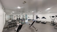Located at 140 Holiday Blvd in Covington, Louisiana, this fitness center offers a well-equipped exercise space designed to support a well-rounded workout routine. The room features a clean, modern aesthetic with bright white walls, recessed and flush-mount lighting, and durable dark rubber flooring that provides excellent traction and comfort underfoot.

The cardio zone is equipped with two treadmills and an elliptical trainer, positioned near a window that allows natural light to filter into the space. For strength training, the facility includes a multi-function cable machine with various attachments, an adjustable weight bench, a dumbbell rack stocked with a range of free weights, and kettlebells — providing ample options for both beginner and experienced exercisers.

Mirror panels along one wall enhance the sense of space and allow users to monitor their form during workouts. A rules and guidelines placard is prominently posted near the entrance, and an intercom system is mounted on the wall for added convenience and safety.

An adjacent laundry area, accessible through a glass-paned door, features stacked washer and dryer units, adding a practical amenity alongside the exercise room. The overall layout is efficient and thoughtfully organized, making the most of the available space while maintaining an open, functional atmosphere.