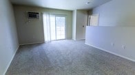 This single-floor space features an open living area finished with neutral wall-to-wall carpeting that extends throughout, contributing to a clean and understated aesthetic. The room is bathed in natural light, courtesy of a large sliding glass door fitted with sheer white curtains that provide privacy while maintaining brightness. The sliding door offers direct access to an outdoor area, allowing for a seamless connection between the interior and exterior. A wall-mounted air conditioning unit is positioned near the sliding door, ensuring climate control within the space. The walls are painted in a soft, muted tone that complements the light carpeting, creating a cohesive and calm atmosphere. A partial wall or half-partition near the entry area subtly defines the transition between the living space and an adjacent area, adding a sense of structure without fully enclosing the layout. The open floor plan allows for flexible furniture arrangement and makes the space feel expansive. Overall, the living area presents a functional and straightforward design, well-suited for comfortable everyday use.