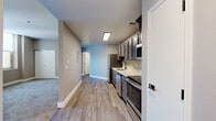 This single-floor unit offers a thoughtfully designed layout that balances functionality and comfort throughout. The kitchen serves as a central highlight, featuring dark-toned cabinetry with ample upper and lower storage, white countertops, and a full suite of stainless steel appliances including a refrigerator, dishwasher, microwave, and range. Light wood-look flooring runs throughout the kitchen and main hallway, lending warmth and continuity to the space. The open corridor connecting the kitchen to the living areas provides a natural flow through the unit. The bedrooms are finished with plush neutral-toned carpeting and benefit from tall windows that invite natural light and offer views of the surrounding urban skyline. Generous closet space is included in the bedroom areas. The bathroom is finished with wood-look flooring consistent with the rest of the unit, featuring a white vanity with a clean countertop surface, a wall-mounted mirror, and matte black hardware accents including a towel bar and toilet paper holder. Neutral warm-gray walls throughout the unit create a calm, cohesive aesthetic. Overall, the space presents a clean, modern interior well-suited for comfortable everyday living.