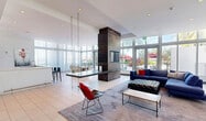 Located at 3635 NE 1st Ave in Miami's vibrant Midtown neighborhood, this single-floor amenity lounge delivers a striking blend of contemporary design and upscale comfort. The expansive open-plan layout is defined by soaring ceilings and walls of floor-to-ceiling glass windows and doors, flooding the space with abundant natural light and offering lush outdoor views of tropical palm-lined surroundings. Sheer white drapery lines the window walls, softening the light and lending an airy, refined atmosphere throughout.

A dramatic double-sided fireplace clad in rich, dark reclaimed wood serves as the architectural centerpiece of the room, anchoring the space and creating a natural division between the lounge and dining areas. A suspended chain-hung table adds an artful, industrial accent overhead. The lounge area features a large sectional sofa, designer wire chairs with bold red cushions, and a tufted ottoman arranged atop an area rug over light-toned tile flooring.

A well-appointed bar area provides a sleek white countertop with seating for multiple guests, set against a full reclaimed wood accent wall housing a mounted flat-screen television and floating white shelves. White cabinetry below provides ample storage. Recessed lighting throughout ensures a warm, welcoming ambiance, making this space ideal for socializing and entertaining.