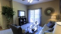 The living area of this single-story space is thoughtfully designed with comfort and functionality in mind. The room features neutral-toned walls that create a warm, inviting atmosphere, complemented by plush carpeted flooring. A ceiling fan with a light fixture is centrally positioned overhead, providing both air circulation and ambient lighting. The focal point of the room is a large sliding glass door framed by sheer patterned curtains, which allows an abundance of natural light to fill the space and provides direct access to a private balcony. The balcony, visible through the glass door, offers outdoor seating and a view of the surrounding area. Inside, the living space is furnished with a sleek sofa and an accent chair with nailhead trim, arranged around a geometric metal coffee table atop a patterned area rug. A dark-framed open shelving unit serves as a media console, adorned with a large mirror above it and small decorative plants. A circular metallic wall accent adds a decorative touch to the opposite wall. The overall ambiance is modern yet cozy, with a balanced mix of practical furnishings and decorative elements.