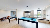 Located at 2 River Plate St in Ajax, Ontario, this contemporary amenity lounge is a well-appointed communal space designed for both relaxation and recreation. The single-floor layout is finished with light hardwood flooring throughout, contributing to a bright and open atmosphere that flows seamlessly across the entire area.

The space is divided into distinct functional zones. A recreation area anchors one side of the room, centered around a full-size pool table with a vivid blue felt surface, set beneath a striking circular pendant light fixture. This zone also includes high-top seating, making it ideal for socializing during gameplay.

On the opposite side, a comfortable lounge area features rich cognac leather armchairs arranged around a low side table, creating an inviting setting for casual conversation. A round dining table with modern chairs occupies an adjacent area, suitable for small group gatherings. A foosball table is also visible, adding to the recreational offerings.

Large windows flood the space with natural light and frame panoramic views of the surrounding Ajax skyline and greenery, enhancing the room's airy feel. The walls are adorned with bold, colorful artwork and framed pieces that lend the space a curated, gallery-like quality. Recessed ceiling lighting complements the natural light throughout. The overall ambiance is modern, vibrant, and thoughtfully designed for community use.