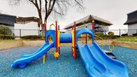 Located at 4333 Chesford Rd in Columbus, Ohio, this outdoor recreational space serves as a dedicated playground area within a residential community. The centerpiece of the space is a colorful, multi-component play structure finished in bold blue, red, and yellow, featuring dual slides, a tunnel connector, climbing steps, and safety railings — designed to engage children of varying ages and abilities. The entire play area is surfaced with blue rubber mulch, providing impact-absorbing cushioning underfoot for added safety. The playground is neatly defined by timber retaining walls that create a raised border around the perimeter, lending the space a structured and well-maintained appearance. Adjacent to the play structure, a spacious gravel courtyard area offers seating via benches and is framed by mature trees, providing a gathering spot for residents. The surrounding residential buildings feature a distinctive architectural design with angled rooflines and open balconies, contributing to the community atmosphere of the grounds. Chain-link fencing encloses portions of the area, offering a secure boundary. The overall setting is an open-air, community-oriented recreational space that combines functional play equipment with thoughtfully landscaped surroundings.