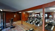 This fitness center is a dedicated exercise facility thoughtfully designed to support a variety of workout routines. The space is defined by its bold, warm-toned walls in deep red and burnt orange hues, creating an energizing atmosphere that motivates physical activity. The flooring features a colorful, patterned hardwood or laminate surface that adds visual interest while providing a durable, easy-to-maintain workout surface. A row of modern cardio machines — including multiple elliptical trainers and steppers — lines one side of the room, positioned in front of a full-length wall mirror that visually expands the space and allows users to monitor their form during exercise. Overhead, flush-mounted fluorescent lighting ensures the room is well-lit throughout. A wall-mounted television is positioned at a comfortable viewing height, offering entertainment during cardio sessions. Structural columns are integrated into the design and finished to match the surrounding walls, maintaining a cohesive aesthetic. A private door at the rear of the room provides additional access or leads to a utility or storage area. The textured ceiling adds a classic architectural finish. Overall, the space is practical, well-organized, and designed to deliver a functional and motivating fitness environment.