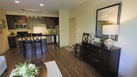 This single-floor residence showcases a thoughtfully designed open-concept layout that connects the living, dining, and kitchen areas into one cohesive and functional space. Rich, dark hardwood flooring runs throughout, providing a warm and sophisticated foundation that ties the entire space together.

The kitchen is a standout feature, outfitted with dark espresso cabinetry, granite countertops, and stainless steel appliances including a refrigerator and dishwasher. A breakfast bar with seating extends from the counter, offering a casual dining option that enhances the open flow of the space.

The dining area is furnished with a dark wood sideboard and coordinating chairs, accented by tasteful décor and a framed landscape artwork that adds a touch of character to the neutral, light-toned walls. Recessed and track lighting are installed throughout, ensuring well-distributed illumination across the space.

The overall ambiance is modern yet inviting, with a neutral color palette of soft greens and whites complementing the dark furnishings and flooring. The layout is efficient and well-organized, making excellent use of the available space while maintaining an open, airy feel. This residence is well-suited for those who appreciate contemporary design with practical, everyday functionality.