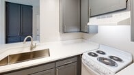 Located at 2235 W Broadway in Anaheim, California, this single-floor unit offers a practical and well-appointed layout designed for comfortable everyday living. The living area is finished with plush wall-to-wall carpeting and neutral-toned walls, creating a warm and inviting atmosphere. A track lighting fixture illuminates the space, while a sliding glass door with vertical blinds opens to an outdoor view, allowing natural light to fill the room. Adjacent to the living area, the kitchen features a peninsula-style counter that serves as both a workspace and a casual divider between rooms. Updated gray cabinetry with brushed nickel hardware lines the kitchen walls, complemented by clean white countertops, a stainless steel sink with a modern faucet, and an electric coil range. Under-cabinet ventilation and ample upper storage add to the kitchen's functionality. The transition between the carpeted living space and the kitchen's hardwood-style flooring provides a clear delineation of areas. A tall built-in storage unit with drawers offers additional organization near the entryway. The bathroom includes a clean, white shower-tub combination with a curved curtain rod. Textured ceilings throughout the unit complete the overall aesthetic of this straightforward and move-in-ready Anaheim residence.