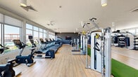Located at 1100 E Apache Blvd in Tempe, Arizona, this modern fitness center offers a generously sized, single-floor layout thoughtfully divided into distinct training zones. The space is bathed in natural light through expansive floor-to-ceiling windows and clerestory windows, offering views of the surrounding Tempe landscape and creating an open, energizing atmosphere throughout.

The cardio zone lines one side of the facility and is outfitted with a full row of treadmills, ellipticals, stair climbers, and stationary bikes — all by Cybex — positioned along the windowed wall to maximize natural light and outdoor views during workouts. Light hardwood-style flooring runs through this area, lending a clean, contemporary feel.

A dedicated functional training zone features a turf strip with sled training equipment, cable machines, a rower, and battle ropes, while full-length mirrors amplify the sense of space. Glass walls provide a visual connection to an adjacent outdoor terrace area.

The free weights and strength zone is outfitted with Hampton dumbbells, barbells, weight plates, kettlebells, a punching bag, and wall-mounted functional rigs. This area features durable epoxy terrazzo flooring, ideal for high-intensity training. A wall-mounted display and bold blue accent tile add visual character to the space. The clean white ceilings with cylinder pendant lighting maintain a bright, modern aesthetic throughout the entire facility.