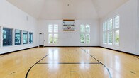 Located at 3055 Riverbirch Dr in Aurora, Illinois, this impressive indoor basketball facility offers a full-size regulation court as its centerpiece. The space is defined by dramatically vaulted, cathedral-style ceilings that rise to striking heights, creating an expansive, open atmosphere ideal for athletic activity. The court itself features a polished hardwood floor with clearly marked regulation lines and key areas, complemented by a wall-mounted adjustable basketball backboard and hoop. Natural light pours in abundantly through large, multi-pane windows positioned along multiple walls and rising to nearly ceiling height, offering views of the surrounding lush greenery. The clean, white-painted walls maintain a bright and energetic environment throughout. An adjacent room — accessible through glass-paneled doors — appears to connect to additional fitness facilities, with basketballs stored on wall-mounted racks nearby, indicating a well-organized, fully equipped athletic space. A fitness area is also visible through an interior window, suggesting the court is part of a broader recreational complex. The overall ambiance is bright, airy, and purpose-built for high-performance sport and recreation, making this an exceptional facility for basketball and other indoor athletic pursuits.