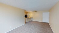 This single-floor unit showcases an open-concept layout that seamlessly connects the living area to the kitchen, creating a spacious and functional flow. The living area is finished with wall-to-wall beige carpeting, contributing to a warm and comfortable atmosphere, while clean white baseboards provide a crisp contrast. Smooth, neutral-toned walls complement the textured popcorn ceiling overhead.

A ceiling fan with a brass-toned fixture is centrally positioned in the living space, providing both air circulation and ambient light. The kitchen is defined by a peninsula-style breakfast bar, which serves as a natural divider between the two areas while maintaining the open feel. Pendant lighting hangs above the bar, adding focused illumination. The kitchen is equipped with a black refrigerator and includes cabinetry visible in the background, along with a sink set within the countertop. White cabinetry and recessed lighting further brighten the cooking space. A white entry door is positioned along one wall of the living area, providing direct access to the unit. Overall, the space presents a clean, move-in-ready aesthetic with practical, comfortable finishes throughout.
