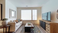 This thoughtfully designed single-floor residence combines modern aesthetics with functional living across a well-organized floor plan. The living room is bright and welcoming, featuring wide windows with horizontal blinds that allow natural light to fill the space, complemented by warm hardwood-style flooring throughout.

The kitchen is a standout feature, showcasing floor-to-ceiling dark cabinetry paired with crisp white countertops and a subway tile backsplash. Stainless steel appliances, including a range, microwave, and refrigerator, add a polished, contemporary feel. A peninsula with seating provides a casual dining option and enhances the open flow between the kitchen and living areas.

The dining area offers an intimate setting with track lighting overhead and direct sightlines to the adjoining rooms, creating a cohesive and connected layout. The bedroom is carpeted for comfort, features a large window for natural light, and includes a ceiling fan for year-round climate control.

The bathroom is finished with large-format tile flooring, dark vanity cabinetry, a dual-sink countertop, and a well-lit mirror, delivering a clean and sophisticated feel. A dedicated in-unit laundry area houses a full-size washer and dryer with overhead shelving for added storage convenience.