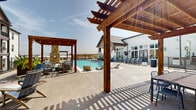 Located at 150 Inspiration Dr in Hudson Oaks, Texas, this outdoor pool area is a well-appointed community amenity space designed for relaxation and recreation. The centerpiece is a generously sized swimming pool with crystal-clear blue water, surrounded by a spacious concrete pool deck lined with contoured lounge chairs, perfect for soaking up the abundant Texas sunshine. Warm wood-beam pergolas provide shaded gathering areas across the deck, adding architectural character and definition to the outdoor space. A standout feature is the stone fireplace lounge area, which offers a cozy, sheltered retreat with comfortable seating for cooler evenings. Adjacent to the pool, a clubhouse-style building with a covered porch, wall-mounted TV, and additional seating areas extends the usability of the space. The surrounding multi-story residential buildings, finished in white and warm wood tones, frame the pool area and contribute to the community's cohesive aesthetic. A separate terrace area with Adirondack chairs offers a quieter vantage point with open views of the surrounding North Texas landscape. The overall ambiance is that of a resort-inspired outdoor retreat, balancing active recreation with comfortable, shaded lounging spaces throughout.