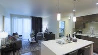 This single-floor residence showcases a thoughtfully designed open-concept layout that seamlessly connects the kitchen, living, and sleeping areas. The kitchen features a sleek white countertop island that serves as both a functional workspace and a social hub, illuminated by elegant pendant lights with cylindrical shades that cast a warm, inviting glow. The open living area is styled with modern furnishings, including a plush area rug over rich hardwood-style flooring, a contemporary accent chair, and a low-profile media console. Large floor-to-ceiling sliding glass doors dressed with dark draping curtains allow abundant natural light to filter in while offering access to an outdoor space. The bedroom, visible through an open passage, is finished with neutral tones and soft furnishings, creating a calm and restful retreat. Throughout the space, a cohesive neutral color palette of whites, grays, and dark accents contributes to a sophisticated and modern ambiance. Recessed lighting complements the pendant fixtures, ensuring the space is well-lit and welcoming at all hours. The overall design balances functionality with contemporary aesthetics, making it a refined and comfortable living environment.
