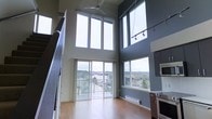 This impressive two-story space is defined by its dramatic double-height ceilings that create an airy, open atmosphere from the moment you step inside. The main living area is flooded with natural light, courtesy of a wall of floor-to-ceiling windows and a sliding glass door that opens to a balcony, framing panoramic views of the surrounding urban landscape and tree canopy beyond. Warm hardwood flooring runs throughout the main level, complementing the bold contrast of dark accent walls against crisp white trim and window frames. A ceiling fan overhead adds a practical touch to the lofty living space, while adjustable track lighting along the upper portion of the wall provides versatile illumination. The kitchen is positioned adjacent to the living area and features stainless steel appliances, including a built-in wall oven and microwave, seamlessly integrating functionality with the modern aesthetic. A staircase with open railings leads to the upper floor, maintaining the sense of openness and visual continuity throughout. The overall ambiance is contemporary and sophisticated, offering a bright, dynamic living environment with an elevated connection to the outdoors.