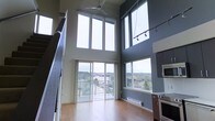 This impressive two-story space is defined by its dramatic double-height ceilings that create an airy, open atmosphere from the moment you step inside. The main living area is flooded with natural light, courtesy of a wall of floor-to-ceiling windows and a sliding glass door that opens to a balcony, framing panoramic views of the surrounding urban landscape and tree canopy beyond. Warm hardwood flooring runs throughout the main level, complementing the bold contrast of dark accent walls against crisp white trim and window frames. A ceiling fan overhead adds a practical touch to the lofty living space, while adjustable track lighting along the upper portion of the wall provides versatile illumination. The kitchen is positioned adjacent to the living area and features stainless steel appliances, including a built-in wall oven and microwave, seamlessly integrating functionality with the modern aesthetic. A staircase with open railings leads to the upper floor, maintaining the sense of openness and visual continuity throughout. The overall ambiance is contemporary and sophisticated, offering a bright, dynamic living environment with an elevated connection to the outdoors.
