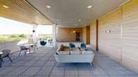 Located at 4501 Park Rd in Charlotte's SouthPark area, this single-floor amenity lounge showcases a contemporary design that seamlessly blends indoor comfort with open-air living. The space is defined by striking horizontal wood-paneled walls that create a warm, natural backdrop throughout, complemented by bold teal accent panels that add a modern pop of color.

The main lounge area is generously proportioned, featuring large-format tile flooring throughout and recessed ceiling lighting that keeps the space bright and welcoming. Multiple seating arrangements are thoughtfully positioned across the floor plan, including a wicker sofa with plush cushions and several rope-woven teal rocking chairs grouped around a round wood coffee table — ideal for social gatherings or relaxed conversation.

The space opens directly to an outdoor terrace, where additional seating and panoramic views of the surrounding Charlotte skyline and greenery are accessible. A corridor with floor-to-ceiling glass-panel doors connects to elevator access and adjacent rooms, including a smaller lounge nook outfitted with a cozy wicker settee. The open-air design and abundance of natural light filtering through the terrace openings give the entire space an airy, resort-like ambiance.