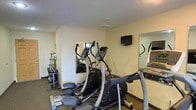 This fitness room provides a dedicated space for exercise and physical wellness. The room features a textured ceiling and neutral-toned walls that create a clean, functional environment. At the center of the space, elliptical machines serve as the primary cardio equipment, offering users a low-impact workout option. A wall-mounted flat-screen television is positioned at eye level, allowing users to stay entertained during workouts. Large wall mirrors are strategically placed to help users monitor their form and technique while exercising, while also contributing to a greater sense of space within the room. A free-weight rack with dumbbells is neatly arranged along one wall, providing options for strength training. The dark carpet flooring adds durability and comfort underfoot. Additional cardio equipment, including what appears to be a treadmill, is visible along the perimeter of the room. The overall layout is efficiently organized to maximize the use of the available space, making it a practical and accessible fitness facility for everyday use.