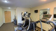This fitness room provides a dedicated space for exercise and physical wellness. The room features a textured ceiling and neutral-toned walls that create a clean, functional environment. At the center of the space, elliptical machines serve as the primary cardio equipment, offering users a low-impact workout option. A wall-mounted flat-screen television is positioned at eye level, allowing users to stay entertained during workouts. Large wall mirrors are strategically placed to help users monitor their form and technique while exercising, while also contributing to a greater sense of space within the room. A free-weight rack with dumbbells is neatly arranged along one wall, providing options for strength training. The dark carpet flooring adds durability and comfort underfoot. Additional cardio equipment, including what appears to be a treadmill, is visible along the perimeter of the room. The overall layout is efficiently organized to maximize the use of the available space, making it a practical and accessible fitness facility for everyday use.