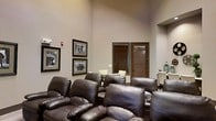 This single-floor space offers a thoughtfully designed common area that combines comfort and style in an inviting setting. The centerpiece of the room is a tiered arrangement of plush, dark leather recliner-style chairs, reminiscent of a private home theater, providing ample seating for gatherings or screenings. The soaring ceilings lend a sense of openness and grandeur to the space, enhancing its overall ambiance. Warm, neutral-toned walls serve as a refined backdrop, accented by a curated selection of framed black-and-white photographs that add a timeless, classic character to the décor. Natural light filters through wood-slatted blinds on a windowed door, contributing a warm and welcoming feel. Adjacent to the main lounge area, a secondary dining or meeting space is visible, featuring light-colored chairs and a table, offering a versatile area for more intimate gatherings or casual use. Architectural details such as a recessed niche and clean wall lines add subtle visual interest throughout. The overall atmosphere is relaxed yet refined, making this space well-suited for entertainment, socializing, or quiet relaxation.