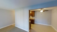 This single-floor space features an open, connected layout that allows the living area to flow naturally into the kitchen. The living area is finished with soft wall-to-wall carpeting in a neutral tone, contributing to a calm and comfortable atmosphere. The walls are painted in a light blue-gray hue, lending the space a clean, airy feel throughout.

A partial dividing wall separates the living space from the kitchen, creating a sense of definition without fully closing off the two areas. The kitchen is equipped with rich, medium-toned wood cabinetry that provides a warm contrast to the lighter surroundings. The countertops feature a granite-look surface, adding a touch of refinement to the functional workspace. A sink is integrated into the counter, and the kitchen floor transitions to a lighter hard-surface material, distinguishing it from the carpeted living area.

A ceiling fan with a light fixture is installed in the kitchen area, offering both ventilation and ambient lighting. White baseboards throughout the space provide clean, finished edges. The overall design is understated and functional, balancing practicality with a welcoming, neutral aesthetic suitable for a variety of uses.