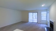 This single-floor residential space offers a clean, straightforward layout designed for comfortable everyday living. The main living area is notably open and airy, finished with wall-to-wall neutral carpeting that adds warmth underfoot. Crisp white walls and a smooth ceiling with recessed lighting contribute to a bright, understated aesthetic throughout the room. A large sliding glass door, dressed with vertical blinds, serves as a focal point and provides direct access to the outdoors, flooding the interior with natural light and offering views of mature surrounding trees. An additional window along the adjacent wall further enhances the sense of openness and ensures the space remains well-lit throughout the day. The overall ambiance is calm and neutral, providing a versatile canvas well-suited for a variety of furnishing styles and personal touches. The single-floor layout ensures easy accessibility and a cohesive flow through the living space. With its combination of natural light, outdoor connectivity, and simple, functional design, this space presents a practical and inviting environment.