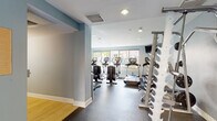 Located at 4041 Radford Ave in Studio City, California, this well-appointed fitness center offers a comprehensive workout facility designed to support a wide range of exercise routines. The space is finished with a durable rubber flooring throughout the main exercise area, complemented by soft blue-gray accent walls and crisp white ceilings with recessed lighting that create a clean, energizing atmosphere.

The cardio zone is a standout feature, lined with large windows that flood the room with natural light and offer views of the surrounding greenery. It is equipped with multiple Life Fitness ellipticals, upright bikes, and treadmills, each featuring individual display screens. Wall-mounted televisions provide additional entertainment options during workouts.

The strength training area is equally well-equipped, featuring a tiered dumbbell rack with a full range of free weights, a multi-function cable machine with various attachments, a weight bench, and a barbell station with plate weights. Floor-to-ceiling mirrors visually expand the space and assist with form.

Adjacent to the fitness room, a tranquil outdoor courtyard is accessible through glass doors and showcases a circular concrete patio surrounded by raised planters filled with tropical foliage and a decorative water feature, offering a peaceful retreat to complement an active lifestyle.