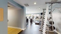 Located at 4041 Radford Ave in Studio City, California, this well-appointed fitness center offers a comprehensive workout facility designed to support a wide range of exercise routines. The space is finished with a durable rubber flooring throughout the main exercise area, complemented by soft blue-gray accent walls and crisp white ceilings with recessed lighting that create a clean, energizing atmosphere.

The cardio zone is a standout feature, lined with large windows that flood the room with natural light and offer views of the surrounding greenery. It is equipped with multiple Life Fitness ellipticals, upright bikes, and treadmills, each featuring individual display screens. Wall-mounted televisions provide additional entertainment options during workouts.

The strength training area is equally well-equipped, featuring a tiered dumbbell rack with a full range of free weights, a multi-function cable machine with various attachments, a weight bench, and a barbell station with plate weights. Floor-to-ceiling mirrors visually expand the space and assist with form.

Adjacent to the fitness room, a tranquil outdoor courtyard is accessible through glass doors and showcases a circular concrete patio surrounded by raised planters filled with tropical foliage and a decorative water feature, offering a peaceful retreat to complement an active lifestyle.
