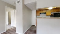 This single-story space is designed with a practical, open layout that connects the main living areas efficiently. The living room serves as a welcoming entry point, featuring wood-look plank flooring, neutral beige walls, and a large window fitted with horizontal blinds that allows natural light to fill the room while offering outdoor views of surrounding greenery. A textured ceiling is consistent throughout the common areas, adding a subtle visual element overhead.

The kitchen is positioned adjacent to the main living space and is separated by a partial wall with a countertop overhang. It is equipped with warm-toned oak cabinetry, a black electric range with a ventilation hood above, a black refrigerator, and a dishwasher. Light-colored countertops provide ample prep space, and the wood-look flooring continues seamlessly from the living area into the kitchen.

A hallway connects the living space to the bedroom and bathroom. The bedroom is generously sized and finished with plush wall-to-wall carpet. Two windows fitted with blinds bring in natural light and views to the exterior. The bathroom includes wood-look flooring, an overhead wood cabinet for storage, and clean white fixtures. A utility access panel is visible in the hallway, and HVAC vents are present throughout for climate control.