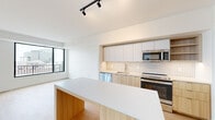 This well-designed one-bedroom, one-bathroom unit offers a thoughtful layout across a single floor, maximizing functionality and flow throughout. The open-concept kitchen and living area serves as the heart of the space, featuring a large island with a wood-paneled base and white quartz countertop, ideal for both meal prep and casual dining. The kitchen is outfitted with stainless steel appliances including a French door refrigerator, range, microwave, and dishwasher, complemented by a herringbone tile backsplash, two-tone cabinetry in white and light wood, and a undermount sink. Light hardwood flooring runs throughout, adding warmth to the clean, modern aesthetic. The living area is bright and airy, with floor-to-ceiling windows framed in matte black that invite natural light and offer open urban views. The bedroom continues the bright, minimalist theme with its own large window and ample square footage. A dedicated walk-in closet provides well-organized storage with built-in shelving and double hanging rods. The hallways are clean and efficiently designed, connecting each area seamlessly. The bathroom features a frameless glass walk-in shower with white subway tile, a wood-vanity with white countertop, and neutral stone-look tile flooring.