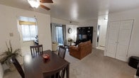 This single-story residence offers an open-concept floor plan that seamlessly connects the dining area and living room, creating a cohesive and functional living space. The dining area is anchored by a dark wood table and matching chairs, accompanied by a ceiling fan overhead that aids in air circulation throughout the room. Natural light filters in through a window positioned near the dining space, brightening the area.

The living room flows directly from the dining area and features plush carpeted flooring in a neutral tone, contributing to the overall warmth of the space. A brown leather sofa serves as the focal point of the seating arrangement, complemented by a cream-colored accent chair nearby. A large, dark entertainment unit lines the far wall, offering ample shelving and storage, with a wall clock displayed above. Sliding glass doors with striped curtains provide access to an outdoor patio area, allowing additional natural light into the space.

A hallway visible from the living room leads to additional rooms, suggesting a practical layout throughout the residence. The white walls and neutral palette create a clean, versatile backdrop throughout.